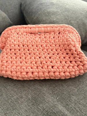 Handvirkad aprikos clutchbag - Söt handvirkad clutchbag i mjukt, tjockt garn i en ljusrosa/aprikos färg. Väskan har en enkel, rektangulär form och stängs med en virkad kant. Perfekt för dig som gillar unika accessoarer och vill sticka ut med något handgjort.