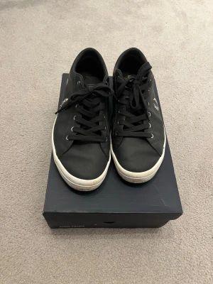 Svarta Fred Perry sneakers - Svarta sneakers från Fred Perry med klassisk låg profil och vita sulor och skolåda ingår. Skorna har svarta skosnören och den ikoniska Fred Perry-loggan i vitt på sidan. Tillverkade i slätt material med enkel och stilren design.