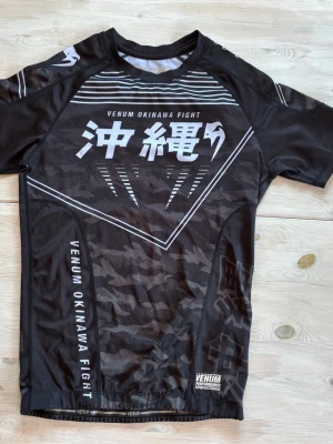 Svart rashguard från Venum Okinawa - Säljer en svart rashguard från Venum Okinawa Fight med korta ärmar. Tröjan har vita och grå grafiska tryck och japanska tecken både fram och bak. Tillverkad i ett stretchigt funktionsmaterial som passar perfekt för kampsport och träning.