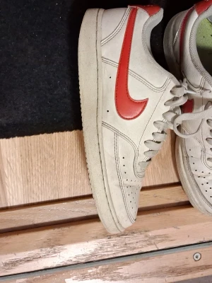 Vita sneakers med rött Nike märke - Snygga vita sneakers från Nike med klassisk röd Nike på sidan. Skorna har perforerad tå för ventilation, vita skosnören och en låg siluett. Tillverkade i läder med detaljer i syntet. Perfekta för dig som gillar retrostil och streetwear.