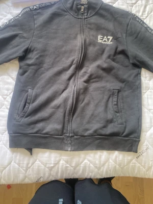 Mörkgrå zip-hoodie från EA7 - Snygg mörkgrå hoodie från EA7 Emporio Armani med dragkedja framtill och två fickor. EA7-logga på bröstet och tryck längs axlarna. Tillverkad i mjukt material som känns skönt mot huden. Perfekt för en avslappnad och trendig look.