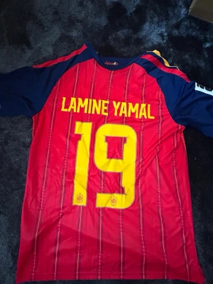Spansk landslagströja Lamine Yamal - Säljer en röd och marinblå fotbollströja från Spaniens landslag med Lamine Yamal och nummer 19 i gult på ryggen. Tröjan har korta ärmar och tunna vertikala ränder i gult och blått. Perfekt för dig som älskar fotboll och vill stötta Spanien.