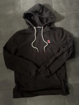 Svart hoodie från Bondelid - Snygg svart hoodie från Bondelid med vit dragsko i huvan och ett rött B-märke broderat på bröstet. Tillverkad i mjukt bomullsmaterial och har långa ärmar samt ribbade muddar. Perfekt för en avslappnad och stilren look.