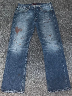 Blå raka jeans från By Identity - Säljer ett par blå jeans från By Identity med raka ben och klassisk femficksmodell. Jeansen har slitningar och unika färgfläckar för en cool, vintage look. Materialet är denim i bomull och passformen är avslappnad.