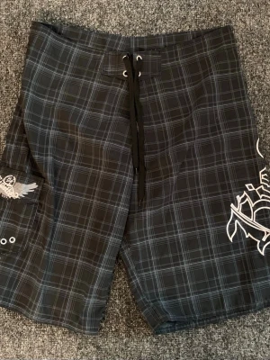 Rutiga svarta badshorts med tryck - Säljer ett par svarta badshorts med vitt rutmönster och coola tryckdetaljer på benet och bak. Shortsen har snörning i midjan och en ficka på sidan. Perfekta för stranden eller poolen, riktigt snygg streetstil.