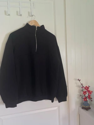 Svart sweatshirt med halv dragkedja - En svart sweatshirt med hög krage och halv dragkedja framtill. Tröjan har långa ärmar och ribbade muddar vid ärmslut och nederkant. Perfekt för en avslappnad och stilren look. Tillverkad i mjukt sweatshirtmaterial.