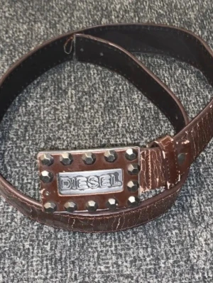 Brunt skärp med nitar från Diesel - Brunt skärp i skinn från Diesel med en stor, rektangulär spänne dekorerad med silverfärgade nitar och Diesel-loggan i mitten. Skärpet har en cool, sliten look och passar perfekt till jeans eller byxor för en edgy stil.
