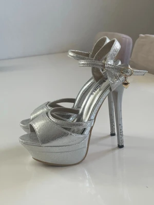 Silvriga platåpumps klackar med strassdetaljer - Snygga silvriga pumps / klackar med hög platåklack, öppen tå och smala remmar runt vristen. Klacken är dekorerad med strass och ett litet hjärthänge i guld hänger vid spännet. Skorna har en glansig metallicfinish och ger en riktigt statement-look.