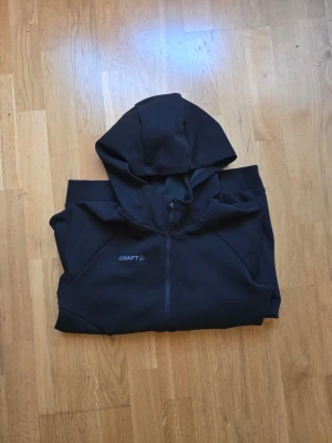 Svart hoodie med dragkedja från Craft - Säljer en svart hoodie från Craft med dragkedja framtill och två sidofickor. Tröjan har huva och ribbade muddar vid ärmsluten. Perfekt för dig som gillar en sportig och enkel stil. Materialet känns mjukt och bekvämt.