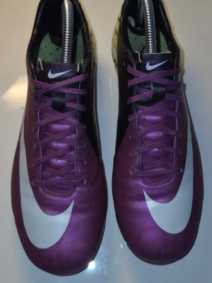 Lila Nike Mercurial fotbollsskor - Säljer ett par lila Nike Mercurial fotbollsskor med stora vita swoosh-loggor på sidan och tå. Skorna har snörning, låg profil och dobbar för spel på gräs. Materialet är syntetiskt och designen är riktigt sportig och modern.