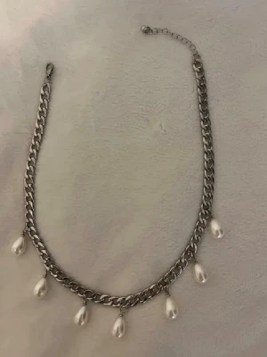 Kedjehalsband med droppformade pärlor - Snyggt halsband med grov silverfärgad kedja och sex droppformade vita pärlor som hänger längs kedjan. Perfekt för att ge en cool touch till din outfit. Justerbar längd med karbinlås. Materialet ser ut att vara metall och konstpärlor.