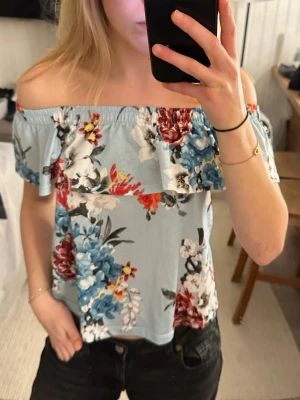 Offshoulder topp  - Säljer en ljusblå offshoulder topp från Gina Tricot i storlek XS. Toppen har ett färgstarkt blommigt mönster i rött, blått, vitt och svart, samt en volang längs axlarna. Perfekt för sommarens! Använd fåtal gånger😊