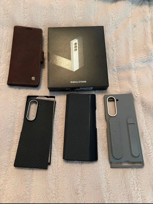 Samsung Galaxy Z Fold6 med tillbehör - Säljer en Samsung Galaxy Z Fold6 med originalkartong och flera fodral i olika material och färger, inklusive svart kolfiberlook, grått plastskal och brunt läderfodral. Telefonen har en stor vikbar skärm och modern design. Perfekt för dig som gillar multitasking och snygg teknik.