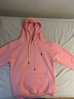 Ljusrosa hoodie - Säljer en ljusrosa hoodie med huva och dragsnören. Hoodien har en klassisk känguruficka framtill och en liten broderad logga på bröstet. Mjuk och skön, perfekt för chill dagar. Passar dig som gillar enkel men snygg stil.
