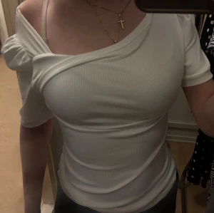 Vit offshoulder ribbad topp - Snygg vit ribbad topp med offshoulder-design och korta puffärmar. Toppen har en omlott-detalj vid bysten och sitter tight. Perfekt för dig som vill ha en trendig och stilren look.