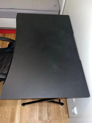 Svart gamingbord med metallställ - Säljer ett svart gamingbord med rektangulär form och lätt böjd framkant. Bordet har ett metallställ på sidan för tillbehör som hörlurar eller kontroller. Bordsskivan är av trä eller kompositmaterial och benen är i metall. Bredden 75cm längden är inte mätt men kan mätas ifall de behövs.