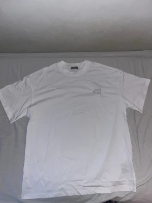 Vit t-shirt  - Säljer en stilren vit t-shirt med diskret broderad logga på bröstet. T-shirten har klassisk passform, rund halsringning och är kortärmad. Perfekt basplagg för alla tillfällen och enkel att matcha med andra plagg.
