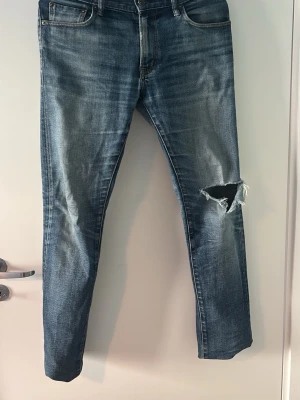 Archive jeans  - Säljer ett par blå jeans med tvättad look och stort hål på ena knät. Jeansen har klassisk femficksmodell och raka ben. Perfekta för dig som gillar en avslappnad och trendig stil.