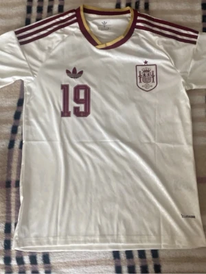 Vit Spanien fotbollströja Adidas - Vit fotbollströja från Adidas med vinröda detaljer, nummer 19 och namnet Lamine Yamal på ryggen. Tröjan har Spaniens landslagsemblem på bröstet, tre ränder på axlarna och gul/vinröd krage. Perfekt för dig som älskar fotboll och vill stötta Spanien.