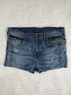True Religion Jeansshorts storlek 34 Blå Stretch - Säljer ett par blå jeansshorts med slitningar och två dragkedjefickor framtill. Shortsen har en klassisk femficksmodell och knappgylf. Materialet är denim med en tvättad look och råa detaljer för en trendig känsla.