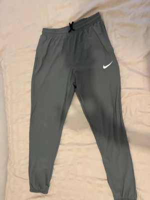 Grå Nike byxor med snörning - Säljer ett par grå mjukisbyxor från Nike med resår och snörning i midjan. Byxorna har smal passform, fickor på sidorna och elastiska benslut. Perfekta för träning eller chill. Nike-logga på ena benet och reflexdetaljer baktill.