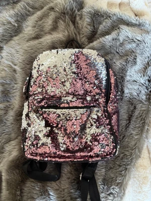 Ryggsäck med rosa och silver paljetter - Glittrig ryggsäck täckt av vändbara paljetter i rosa och silver. Väskan har en framficka med dragkedja och svarta justerbara axelband. 