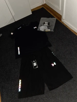Svart Under Armour set med logga - Säljer ett svart set från Under Armour med t-shirt och shorts. Båda plaggen har reflekterande loggor och textdetaljer i regnbågsfärger. Setet är gjort i ett mjukt och lätt material, perfekt för en sportig och avslappnad stil.