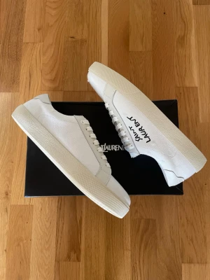 Vita Saint Laurent canvas sneakers - Snygga vita sneakers från Saint Laurent i canvas med krämfärgade sulor och vita skosnören. Klassisk låg modell med svart Saint Laurent-tryck på sidan. Perfekta för en clean och stilren look.