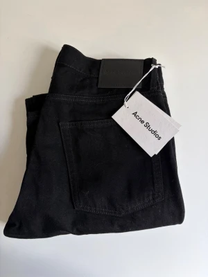 Svarta jeans från Acne Studios - Snygga svarta jeans från Acne Studios med raka ben och klassisk femficksmodell. Jeansen har en stilren look med diskret logga på bakfickan och är tillverkade i ett kraftigt bomullstyg. Perfekta för dig som gillar minimalistisk och trendig stil. Aldrig använda!