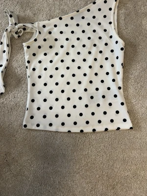 Polka dot topp med knyt - Snygg topp med svarta prickar och knyt vid axeln. Toppen har en tight passform och är perfekt för dig som vill sticka ut lite extra. Materialet känns mjukt och stretchigt. Priset kan diskuteras!
