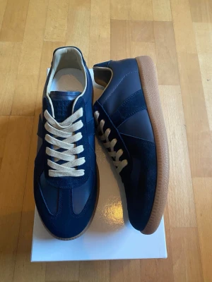 Mörkblå Margiela Gats med beige sula - (Helt Nya )Snygga mörkblå sneakers med ovandel i mocka och slät textil, beigea skosnören och klassisk gummisula i ljusbrunt. Stilren design med diskreta paneler och låg profil som passar till många outfits.