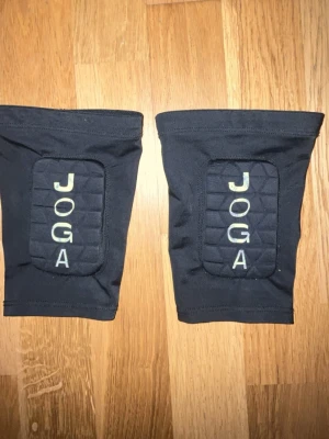 Svarta knäskydd från JOGA - Säljer ett par svarta knäskydd från JOGA med vadderade partier framtill för extra skydd vid träning eller sport. De är elastiska och sitter tajt runt knäna, perfekta för dig som vill ha stöd och komfort under aktivitet.