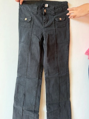 Svarta jeans från DIVIDED, strl 34 - Säljer ett par svarta jeans från DIVIDED i storlek 34. Byxorna har raka ben, två fickor med lock och knapp framtill samt bälteshällor. Materialet är mjukt och har en klassisk denimlook. Perfekta för en trendig och avslappnad stil.