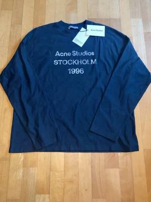 Mörkblå långärmad topp Acne Studios - Snygg mörkblå långärmad topp från Acne Studios med tryck 'STOCKHOLM 1996' framtill. Klassisk rak passform och rund halsringning. Perfekt för dig som gillar stilrena och trendiga plagg med en urban touch.