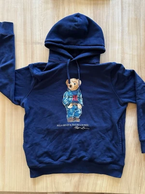 Ralph Lauren Marinblå hoodie - Snygg marinblå hoodie från Ralph Lauren med det ikoniska Polo Bear-trycket på framsidan. Tröjan har huva med dragsko och ribbade muddar. Tillverkad i mjukt bomullsmaterial som är skönt mot huden. Perfekt för en avslappnad och trendig look.