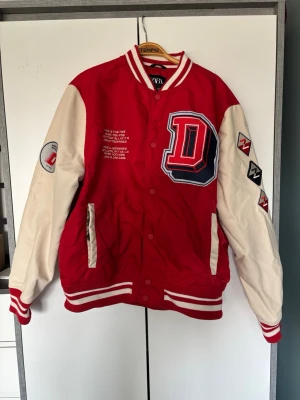 Röd och beige bomberjacka från ZARA - Plagget är bara skrynkligt, inte förstört. Pris går att diskutera. Jag fick jackan av en bekant, jag har inte använt den. Jag är 175 för referens. Snygg röd bomberjacka från ZARA med beigea ärmar och coola patches på båda ärmarna. Stor D-applikation på bröstet och vit text på framsidan. Jackan har ribbade muddar i rött och vitt samt två fickor framtill. Perfekt för en sportig och trendig look. 
