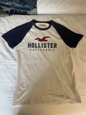 Vit och marinblå Hollister t-shirt - Säljer en klassisk t-shirt från Hollister med vit kropp och marinblå ärmar. Stor logga och text på bröstet i mörkblått och rött. Skön passform och perfekt till vardags.