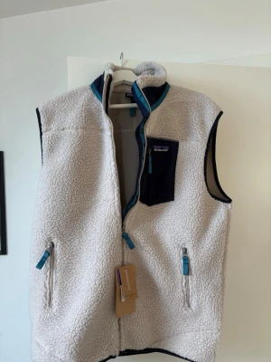 Vit retro fleeceväst från Patagonia - Snygg vit fleeceväst från Patagonia med blå detaljer och dragkedjor. Västen har en hög krage, två sidofickor med dragkedja och en bröstficka i marinblått tyg. Perfekt för lager-på-lager och chill stil.