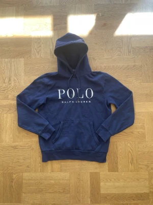 Ralph Lauren hoodie  - Snygg marinblå hoodie från Polo Ralph Lauren med stor vit logga på bröstet och broderad häst på ärmen. Klassisk känguruficka framtill och justerbar huva med dragsko. Tillverkad i mjuk bomullsblandning för skön känsla. 