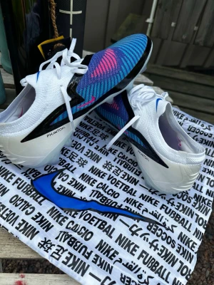 Nike Phantom blå/vita fotbollsskor - Säljer ett par Nike Phantom fotbollsskor i vitt och blått med svarta och rosa detaljer. Skorna har en stickad ovandel med mönstrade ränder och vita snören. Sulan är vit och skon har en modern, strömlinjeformad design perfekt för snabba spelare.