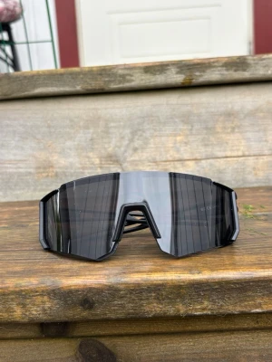 Svarta sportiga solglasögon - Säljer ett par riktigt snygga, sportiga solglasögon med svart, kantig båge och stora, mörka linser. De har en futuristisk vibe och passar perfekt för dig som vill sticka ut. Glasögonen har en bred, rektangulär form och ger ett coolt intryck.