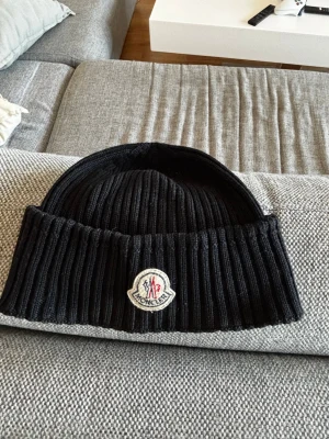 Svart ribbstickad mössa från Moncler - Säljer en svart ribbstickad mössa från Moncler med uppvikt kant och klassiska Moncler-loggan framtill. Mössan är tillverkad i ett mjukt och stretchigt material som sitter skönt på huvudet. Perfekt för dig som vill ha en stilren och exklusiv accessoar.