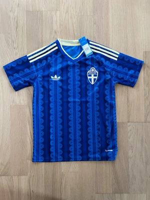 Blå Sverige fotbollströja Adidas - Blå fotbollströja från Adidas med Sveriges landslagsemblem på bröstet. Tröjan har ett vågigt mönster, gula detaljer och tre klassiska ränder på axlarna. Tillverkad i lätt och ventilerande material, perfekt för träning eller match.