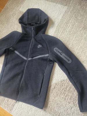 Nike Tech Fleece hoodie svart - Nike Tech Fleece hoodie i svart med hel dragkedja, huva och diskreta grå detaljer. Snygg design med fickor och en extra ficka med dragkedja på ärmen. Perfekt för en sportig och avslappnad stil. Storleken är Small alltså att det är de den passar. Helt ny nike tech, samma mjuka sköna och mörka material som den kom med, inget är dåligt tvättat, allt i bra sick. 