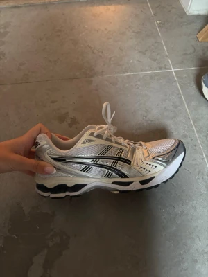 Asics gel kayano 14 - Snygga sneakers från Asics i vitt och silver med svarta detaljer. Skorna har en sportig design med meshpaneler för bra ventilation och klassiska snörningar. Perfekta för dig som gillar retrostil och bekväma skor till vardags.