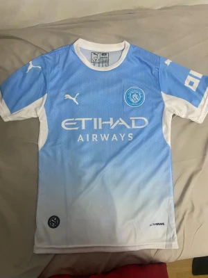 Manchester City matchtröja Puma - Snygg Manchester City matchtröja från Puma i ljusblått och vitt med klubbmärke på bröstet och Etihad Airways-tryck. Tröjan har korta ärmar, rund hals och är tillverkad i ett lätt och ventilerande material perfekt för fotbollsträning.