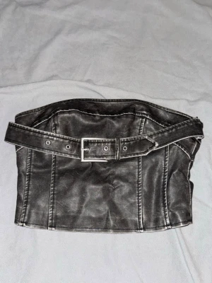 Svart topp med bälte från Soya - Snygg svart korsett från Soya Fashion i konstläder med markerade sömmar och ett brett bälte med metallspänne. Dragkedja bak och fodrad insida för extra komfort. 