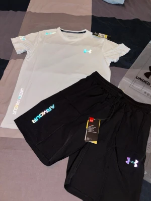 Under Armour träningsset t-shirt & shorts - Säljer två snygga träningsset från Under Armour. Ena setet har vit t-shirt och svarta shorts, det andra har svart t-shirt och svarta shorts. Båda seten har reflekterande loggor och text, samt elastisk midja på shortsen. Perfekt för gym eller löpning.