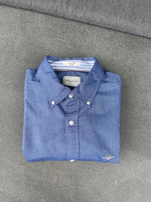 Blå skjorta från Gant i bomull - Klassisk blå skjorta från Gant i mjuk bomull. Skjortan har button-down krage, vita knappar och en liten broderad logga vid nederkanten. Insidan av kragen har ett randigt mönster i vitt och blått för en extra detalj.
