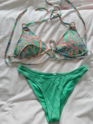  🌴 Grön bikini med guldringar & ribbad textur (Stl M/L) - Drömmig grön bikini som aldrig är använd! Säljes då den tyvärr inte kommer till användning.Mönstrad triangelöverdel med justerbar knytning + guldringar ✨Enfärgad, ribbad underdel med ett jättefint subtilt glans.Snabbtorkande material av hög kvalitet.Passar dig som har storlek M eller L. Skriv i chatten om du vill ha fler bilder! ☀️